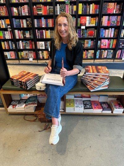 Photo of a woman sitting in a bookshop with stacks of books, signing a book